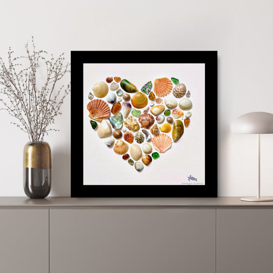 Heart-shaped sea glass and shell mosaic by Steffi Tregenna in soft greens, peach, and blues, framed in pale wood, titled NZ Sea Treasures Heart III sitting on a side shelf with lamp and dried florals