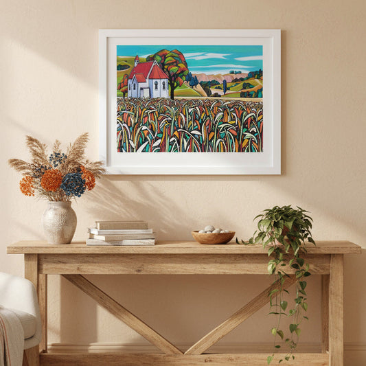 Framed artwork of Waimarama Church by Helena Verhaaren, above a wooden console table in a room with a chair and decorative items.