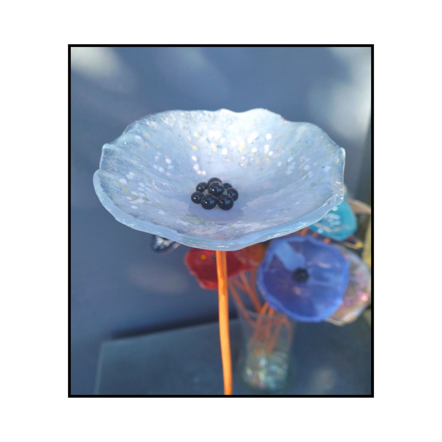 Close-up of a soft pale blue glass flower with a dark centre and bent copper stem