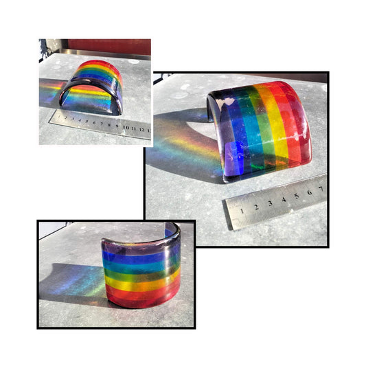 Small fused glass rainbow with reflection sitting on a metal surface with bush behind and a ruler for size comparison