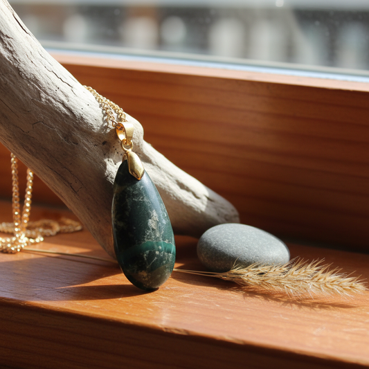 Greenstone teardrop pendant by Gary Howse draped over driftwood on a warm NZ timber windowsill with soft natural light and dried native grass