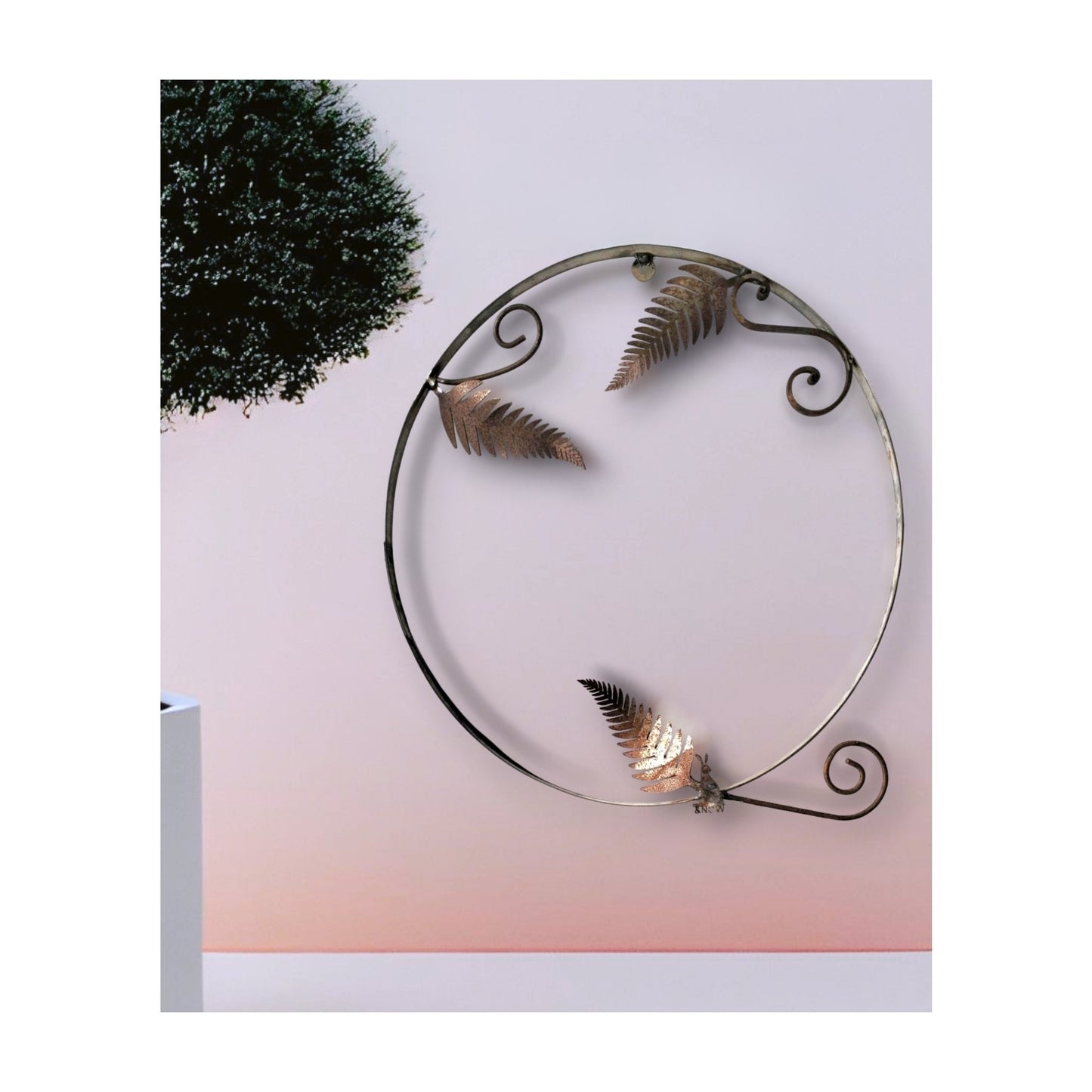 Large circular metal wall sculpture with laser-cut fern and hare patterns casting shadows, displayed on a white wall with a potted plant nearby