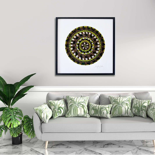 Framed mandala art made from Feathers above a gray sofa with green patterned cushions in a living room setting.