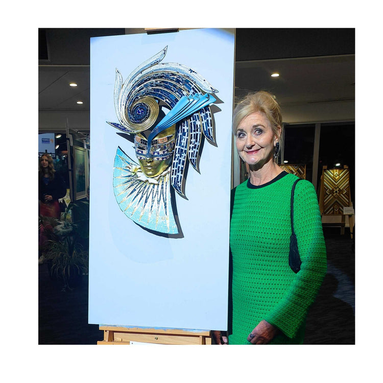 Artist Janice Corbishley standing next to one of her mosaic mask pieces at an exhibiton