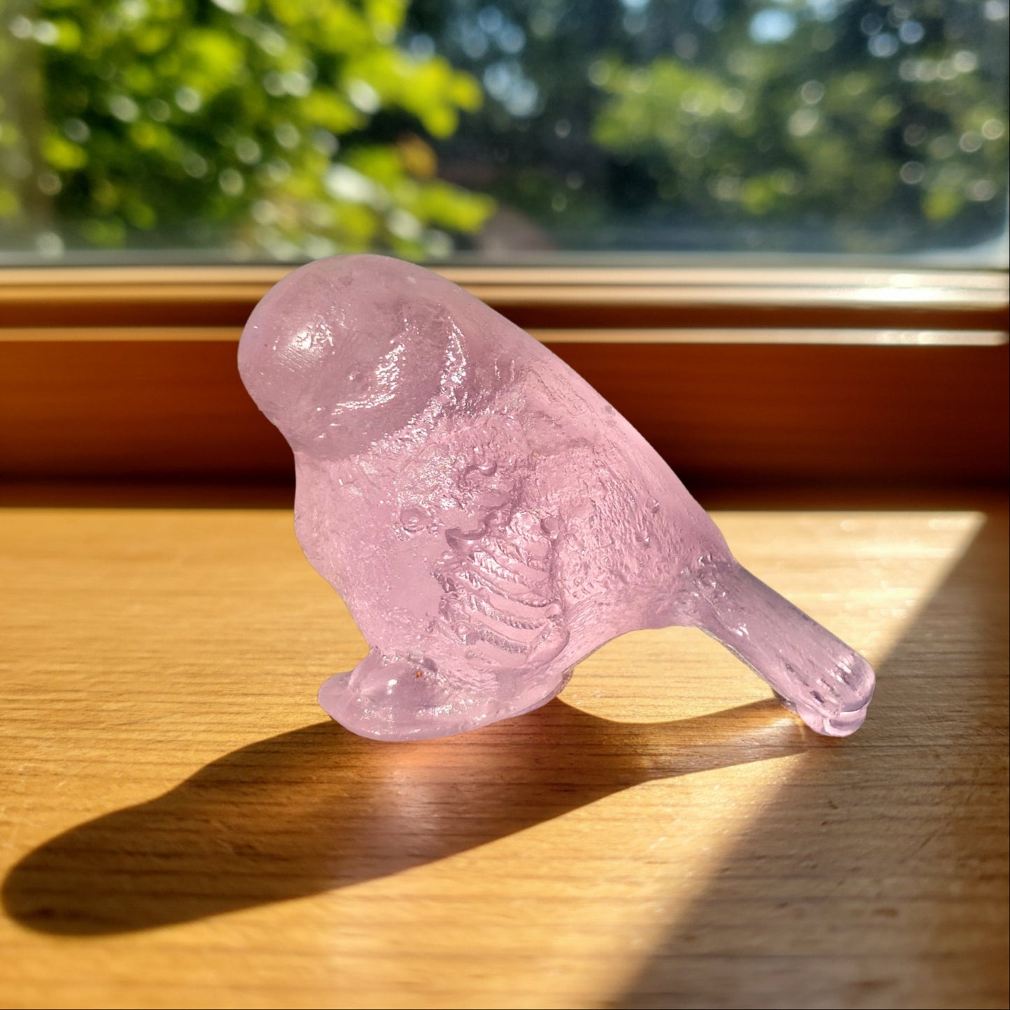 Pink cast glass bird sculpture by Lou Mathieson glowing on a sun-drenched timber windowsill with rosy light reflection
