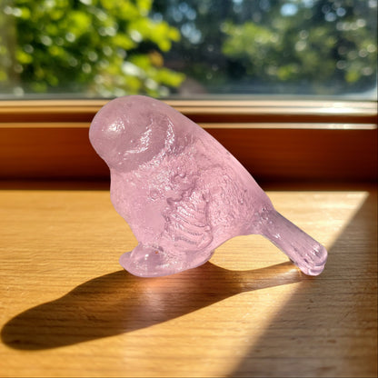 Pink cast glass bird sculpture by Lou Mathieson glowing on a sun-drenched timber windowsill with rosy light reflection

