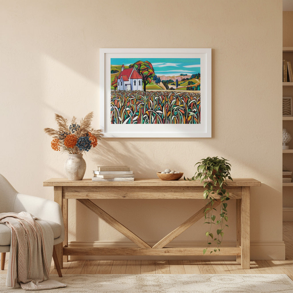 Framed artwork  of Waimarama Church by Helena Verhaaren, above a wooden console table in a room with a chair and decorative items.