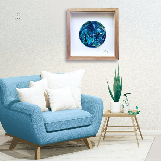 Blue armchair with white cushions in a room with a framed beaded blue embroidery artwork on the wall.