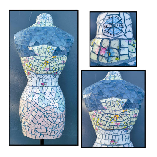 A mosaic bust mannequin torso shown from the front and back in two inset views, covered in blue, white, and pale pink tile-like pieces with small multicolor accents. A circular logo reading 'colchester mosaic' is visible on the back artist Janice Corbishley