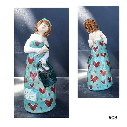 Mandy Olson’s ceramic lady 03, with brown hair, a blue dress with red heart shape pattern, she is holding a wine glass and a bag with two bottles of wine, shown from the front and back.