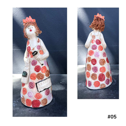 Mandy Olson’s ceramic lady 57, with reddy brown hair, a white dress with colourful koru shape pattern, she is holding a wine glass and a bottle of wine, shown from the front and back.