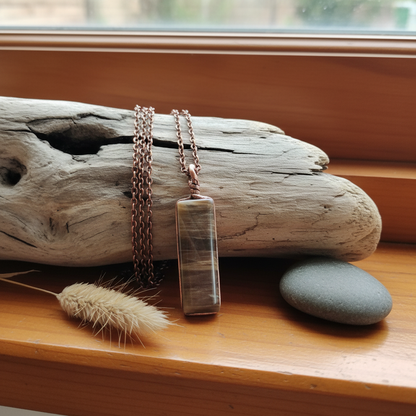 Stripped brown stone pendant by Gary Howse draped over driftwood on a warm NZ timber windowsill with dried native grass and river stone
