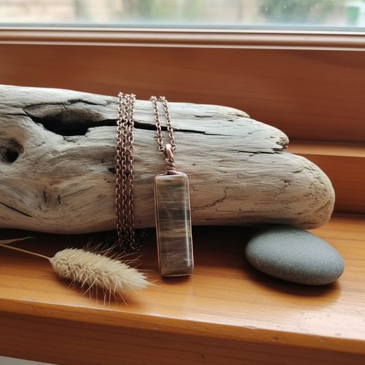Stripped brown stone pendant by Gary Howse draped over driftwood on a warm NZ timber windowsill with dried native grass and river stone
