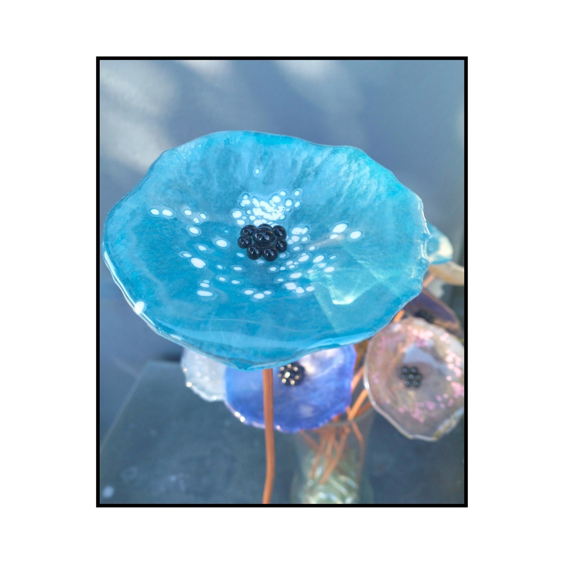 Close-up of abright blue glass flower with white dots with a dark centre and bent copper stem