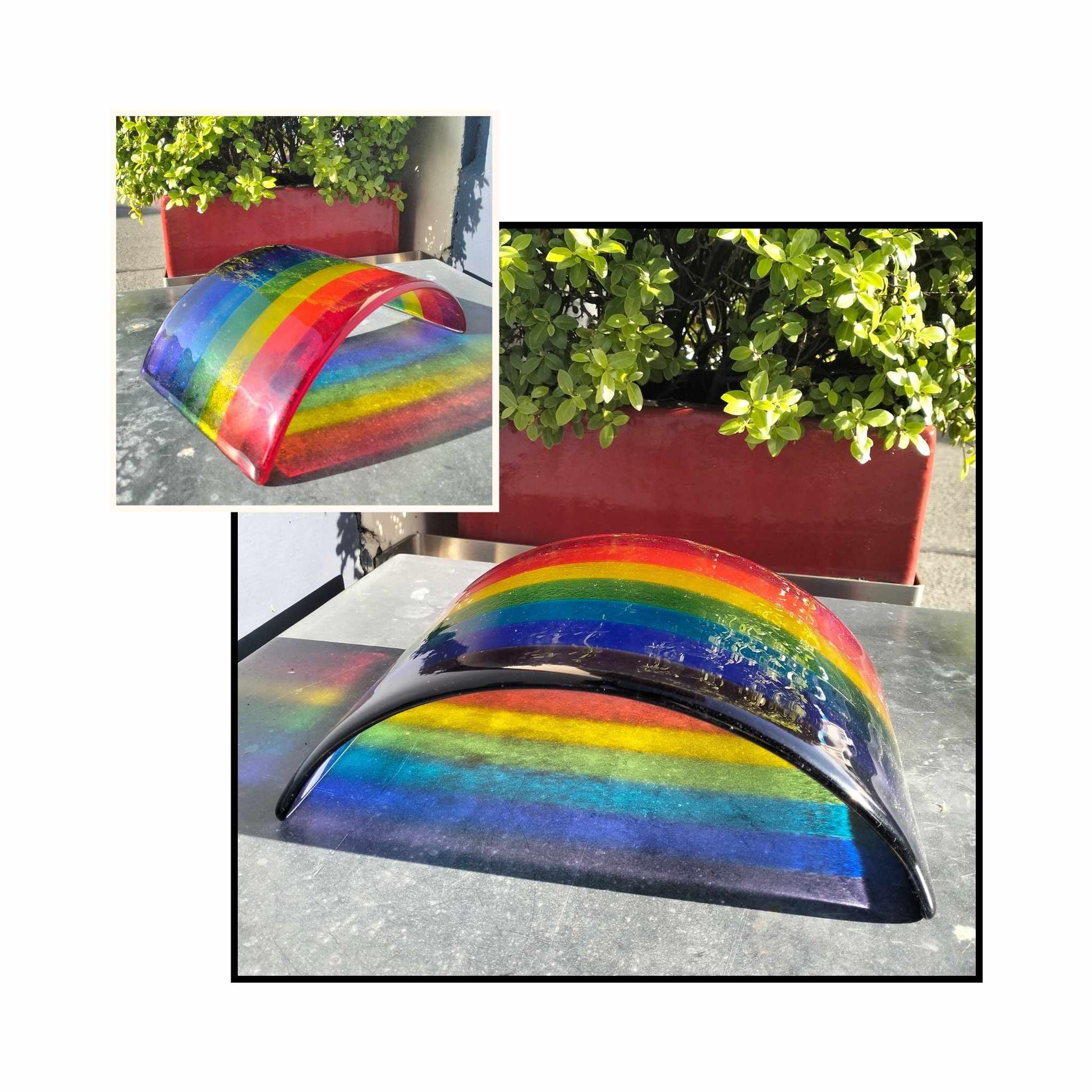 Large fused glass rainbow with reflection sitting on a metal surface with bush behind.