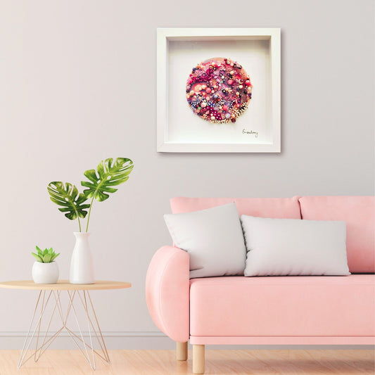 Framed artwork of soft pink beaded embroidery above a pink sofa with a small table and plants in a room