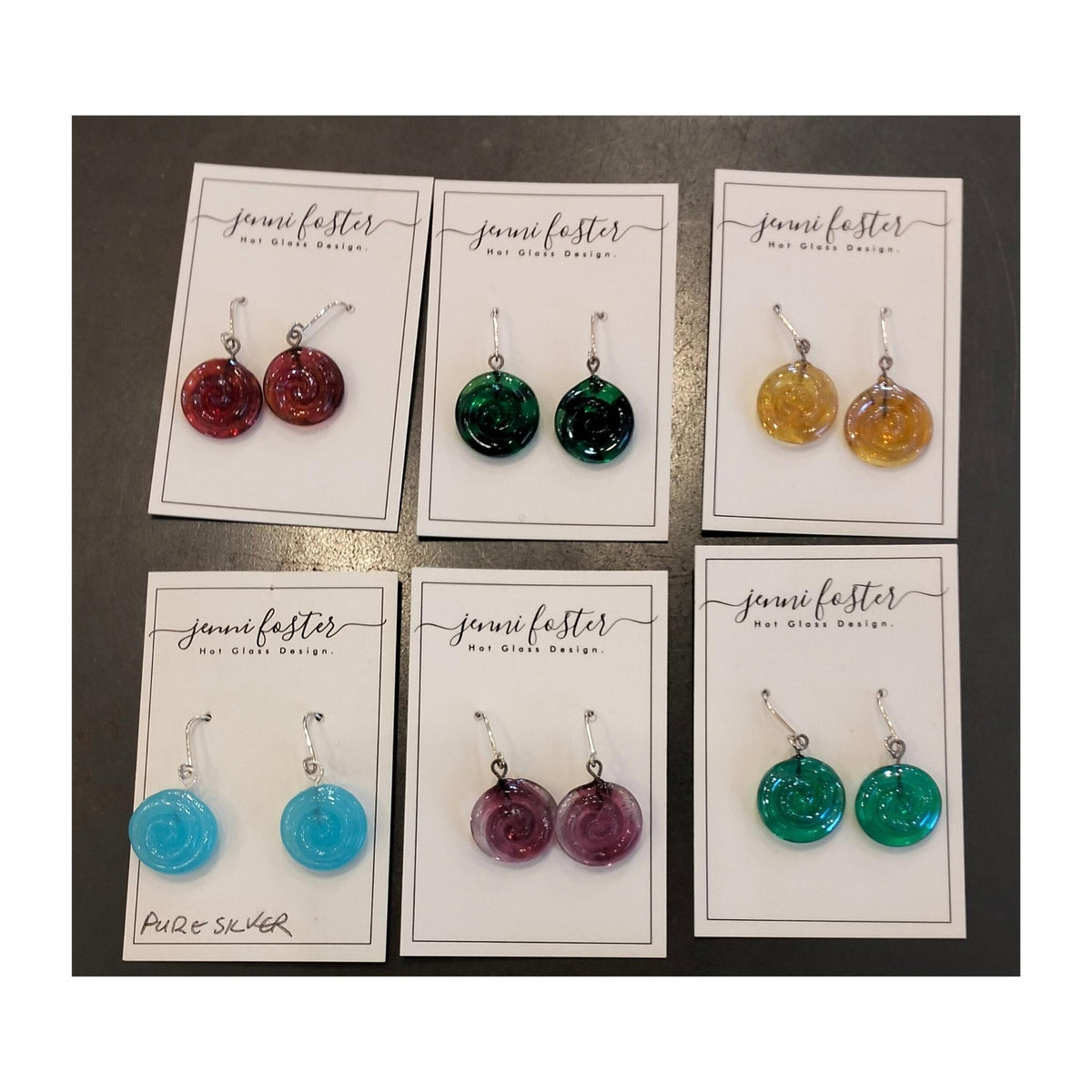 Six pairs of Jenni Foster’s round glass koru earrings in red, dark green, amber, light blue, purple and turquoise, displayed on jewellery cards.
