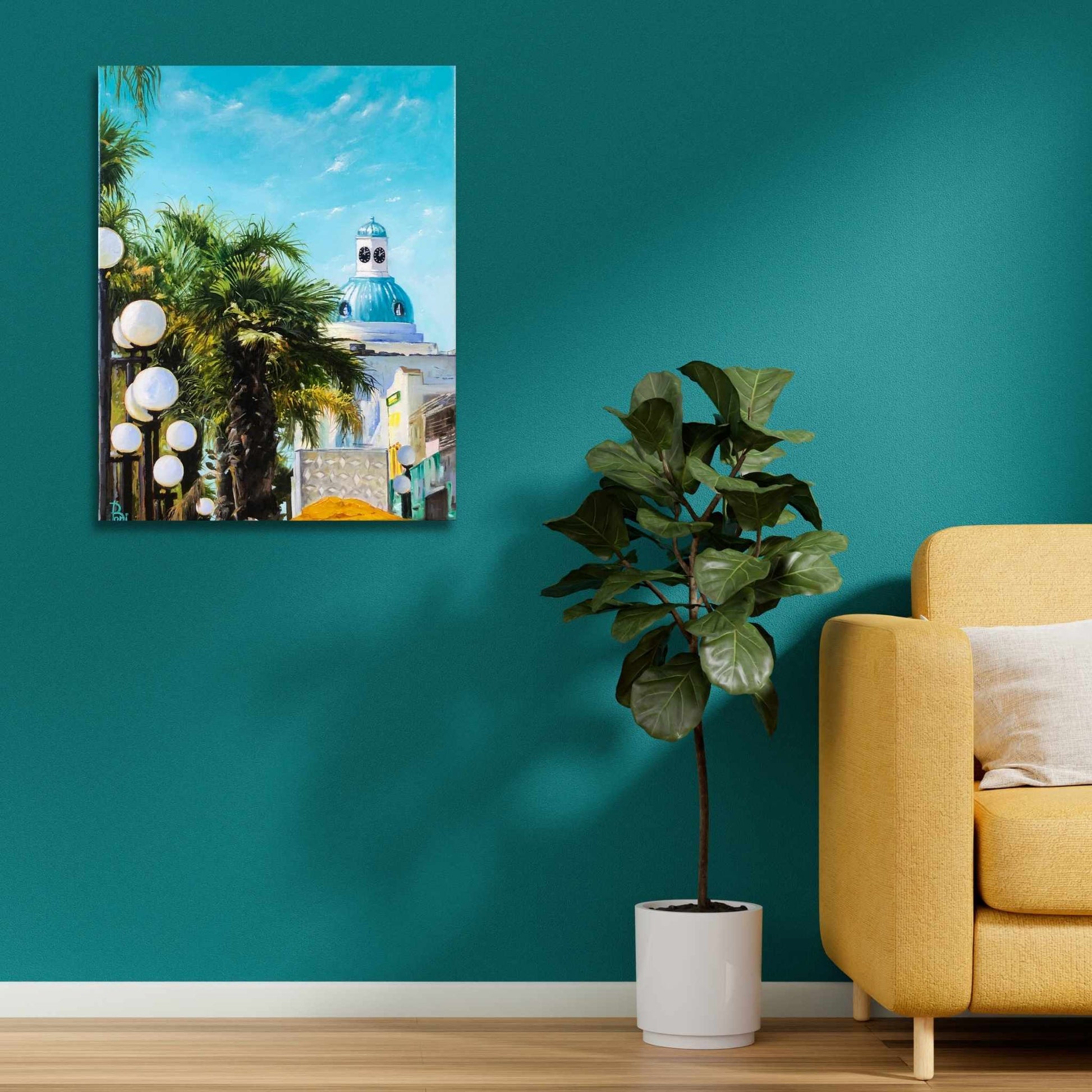 Living room with teal wall, yellow sofa, potted plant, and framed artwork of Painting of sunny Napeir the Art Deco capital of the world.  Scene includes  palm trees, street lights, and a building with a blue dome with a clock tower.